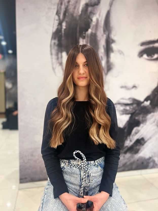Woman with long wavy hair, wearing black top and jeans, seated in front of a monochrome mural.
